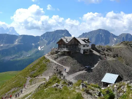 Rifugio alpino su un crinale roccioso, con sentiero tortuoso e escursionisti. Un'auto si avvicina al passo stalle.