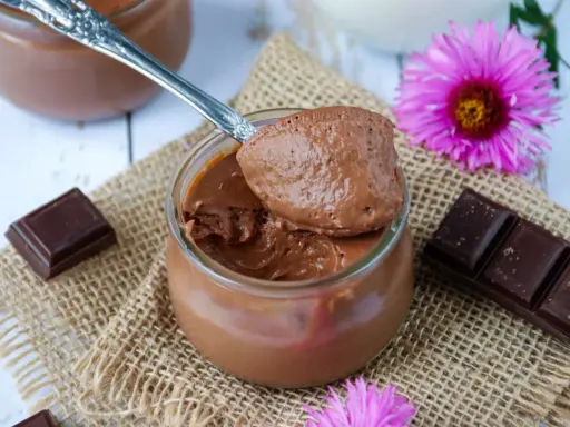 Une cuillère de crème au chocolat sans sucre, onctueuse et aérienne, sur un pot en verre. Des carrés de chocolat et des fleurs roses décorent la scène.