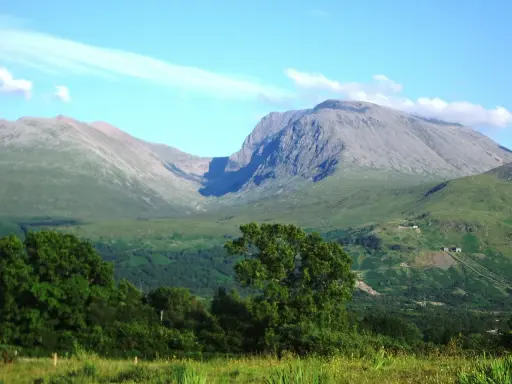 Majestatyczny Ben Nevis wznosi się na tle błękitnego nieba z delikatnymi chmurami. Zielone zbocza górskie i bujna roślinność tworzą malowniczy krajobraz.