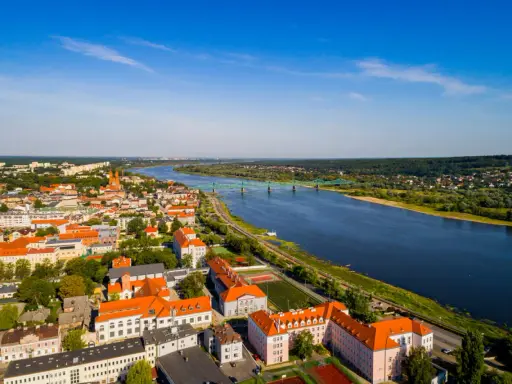 Widok z lotu ptaka na miasto nad Wisłą, z zielonym mostem i zabudowaniami w słoneczny dzień.