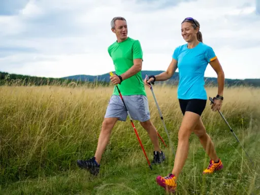 Ein Paar genießt das **power walking** in einer sonnigen Landschaft mit hohem Gras und bewaldeten Hügeln im Hintergrund.