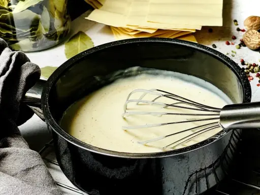 Cremige Béchamelsauce wird in einem Topf mit einem Schneebesen zubereitet. Daneben liegen Lasagneplatten und Gewürze.