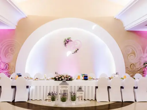 Elegancka sala weselna z białymi pokrowcami na krzesłach i serwowanym stołem. Serce z imionami pary młodej zdobi ścianę, tworząc atmosferę wesela z klasą.
