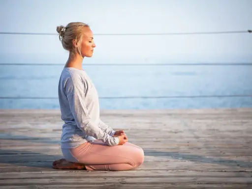 Kvinna med slutna ögon sitter i meditationsställning på en träbrygga vid havet, utövar progressiv avslappning.