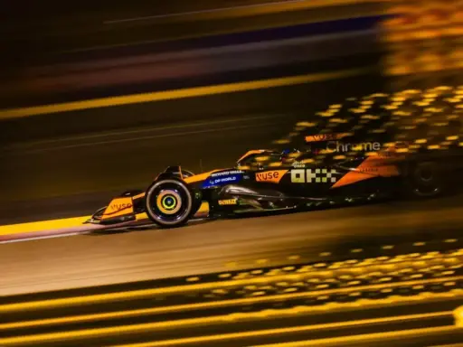 Un coche de **fórmula 1** naranja y negro acelera en la noche, dejando estelas de luz dorada.