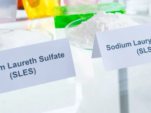 Zwei Schalen mit weißen Pulvern, beschriftet mit "Sodium Laureth Sulfate (SLES)" und "Sodium Lauryl Sulfate (SLS)". Diese Inhaltsstoffe sind oft in SLS-Shampoos zu finden.