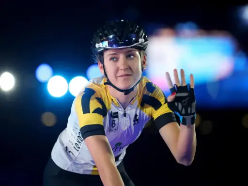 Lea Lin Teutenberg winkt mit der Hand, trägt Radsportkleidung und einen Helm. Blaue und weiße Lichter im Hintergrund.
