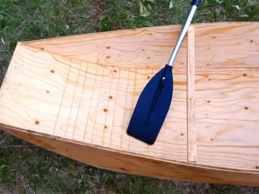 Bateau en contreplaqué brut, prêt pour la résine époxy. Une pagaie noire repose à l'intérieur.