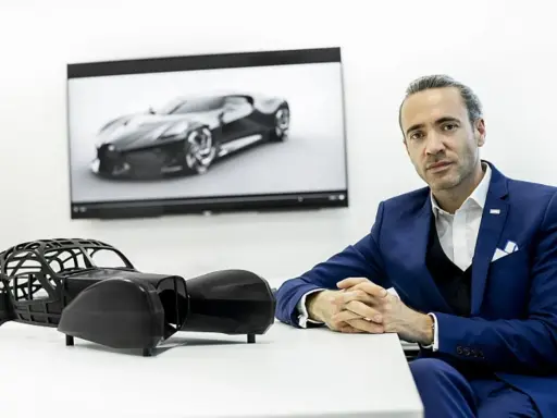 Ein Mann in einem blauen Anzug sitzt vor einem Bildschirm mit dem Bugatti La Voiture Noire. Daneben ein Modell des Autos.