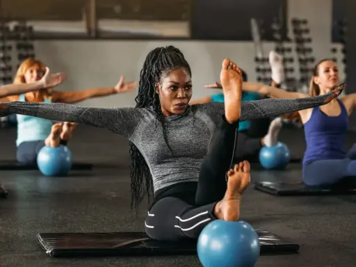 Frauen in einem Kurs, der Pilates Krafttraining macht, sitzen auf Matten mit kleinen blauen Bällen unter ihren Füßen.
