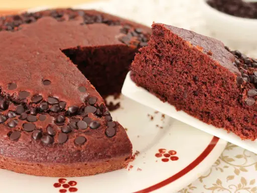 Una fetta di torta al cioccolato morbida, cosparsa di gocce di cioccolato, viene servita da una tortiera intera.