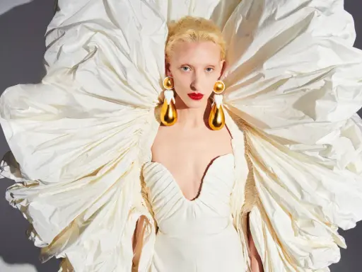 A model wears a dramatic white gown with a voluminous, ruffled cape, showcasing Daniel Roseberry's avant-garde design and striking gold teardrop earrings.