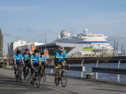Fünf Radfahrer in blauen Trikots trainieren für einen Mini-Triathlon am Hafen, ein großes Kreuzfahrtschiff im Hintergrund.