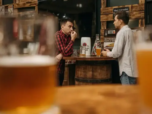 Dwóch mężczyzn rozmawia przy barze, zastanawiając się, ile gram alkoholu ma piwo. Na blacie stoją kufle i piwo.