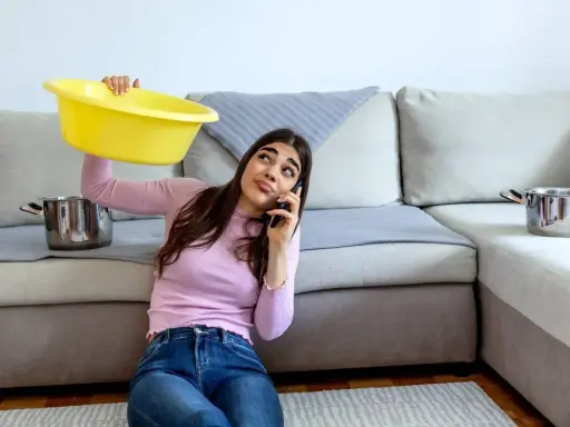 Mujer preocupada llama por teléfono mientras sostiene un barreño amarillo para recoger agua de las goteras en casa. Ollas esperan en el sofá.