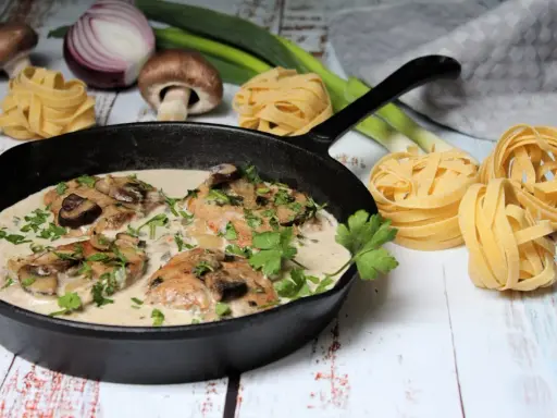 Ein köstliches Schnitzelpfanne Rezept mit cremiger Pilzsoße in einer Gusseisenpfanne, garniert mit Petersilie. Daneben liegen frische Pasta-Nester, Pilze und Lauch.