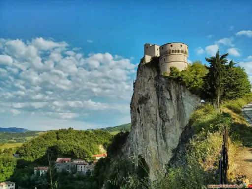 La fortezza di San Leo domina un picco roccioso, con un borgo sottostante e un cielo azzurro punteggiato di nuvole.