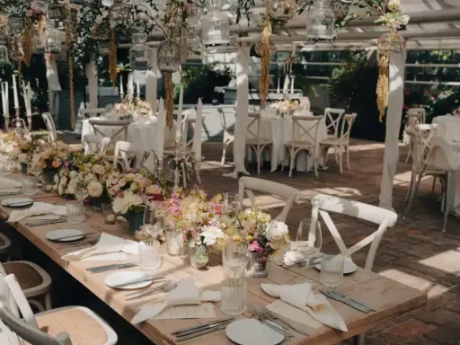 Romantische Hochzeitslocation Ideen: Ein langer Holztisch mit Blumenarrangements, Kerzen und weißen Stühlen, bereit für Gäste.