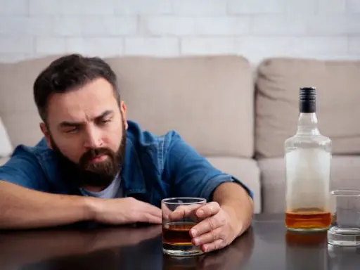 Mężczyzna z zarostem patrzy na szklankę z alkoholem, widoczne objawy uzależnienia. Obok pusta butelka i kolejna szklanka.