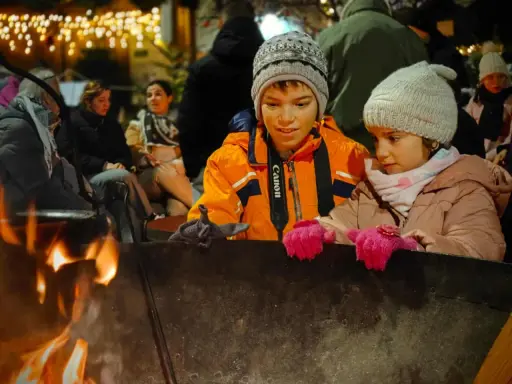 Niños disfrutan de una fogata en Italia, con luces festivas de fondo.