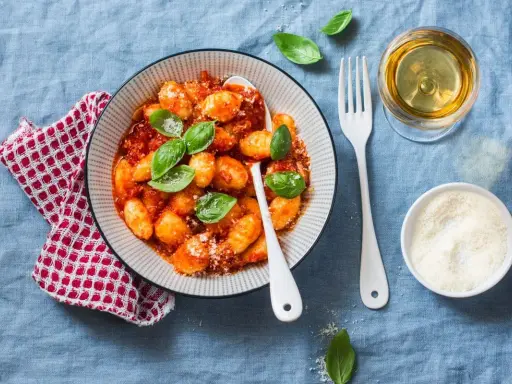 Pyszne kluski gnocchi w sosie pomidorowym, udekorowane bazylią. Obok kieliszek białego wina i miseczka parmezanu.