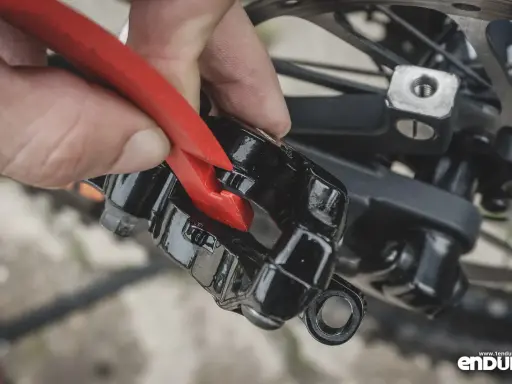 Dłoń z czerwonym narzędziem wykonuje regulację hamulca tarczowego w rowerze, przybliżając klocki do tarczy.