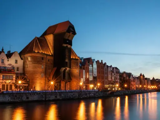 Gdańsk nocą. Historyczny Żuraw i kamienice nad Motławą, rozświetlone latarniami, odbijają się w wodzie.