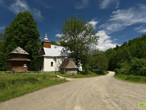 Cerkiew w Łopience, drewniana chata i kamienna przybudówka na tle zieleni i błękitnego nieba z chmurami.