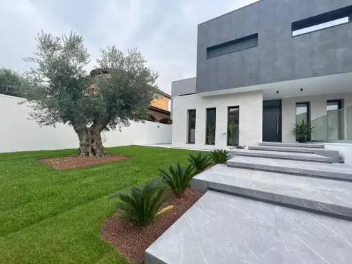 Moderna casa con fachadas color gris y blanco, escaleras de mármol y un olivo centenario en el jardín.