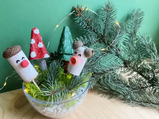 Uroczy stroik DIY z korkowych ludzików, grzybków i choinek w szklanej misie, ozdobiony gałązkami świerku i lampkami.