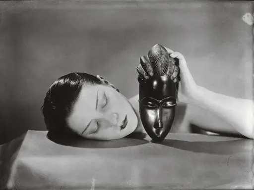 Foto Man Ray: volto femminile addormentato accanto a una maschera africana tenuta da una mano.