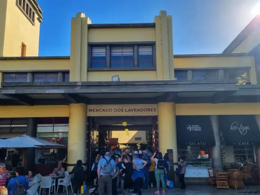 Menschen drängen sich vor dem Mercado dos Lavradores, einem gelben Gebäude mit Cafés und Geschäften.