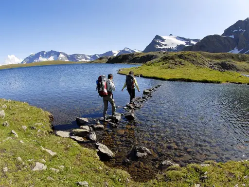 Due escursionisti attraversano il lago di Ceresole su un sentiero di pietre, con montagne innevate sullo sfondo.