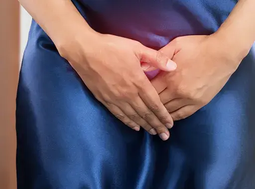Kobieta w granatowej sukience z rękami na podbrzuszu, w tle toaleta. Problem z nietrzymaniem moczu? Czas na wizytę u lekarza.