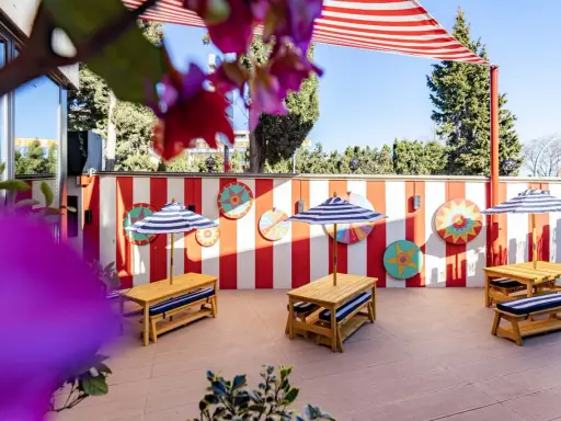 Terraza de restaurantes con zona infantil en Madrid, decorada con rayas rojas y blancas y coloridos adornos circulares. Mesas de madera con sombrillas a rayas invitan a disfrutar.