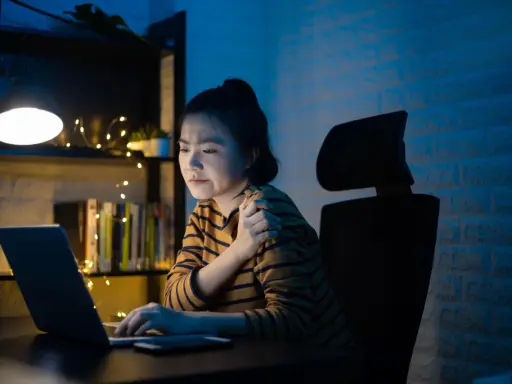 Une jeune femme, en pleine **mise en place télétravail**, ressent une douleur à l'épaule devant son ordinateur, éclairée par une lampe de bureau.