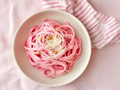 En skål med rosa pasta, toppad med pinjenötter och riven ost. En randig duk i rosa och vitt ligger bredvid.