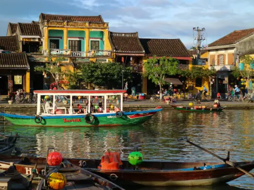Båtar flyter på floden i Hoi An, Vietnam, med färgglada hus och folk som promenerar längs kajen.