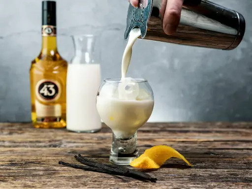 En hand häller en krämig dryck från en shaker i ett glas med is. Bredvid står en flaska Licor 43, en karaff med mjölk, vaniljstänger och citronskal.