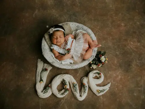 Dolce foto neonato 1 mese dorme in una cesta, circondato da fiori e dalla parola "love" creata con tessuto.