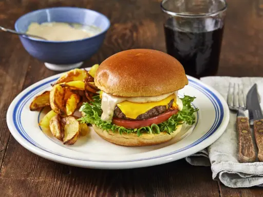 En saftig hamburgare med ost, sallad och tomat, serverad med klyftpotatis och en skål med hemlagad hamburgerdressing.