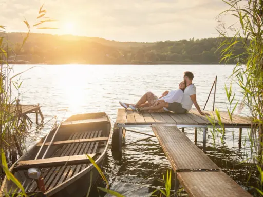 Para, gdzie na Mazury we dwoje? Para siedzi na pomoście, łódka przy brzegu, zachód słońca.
