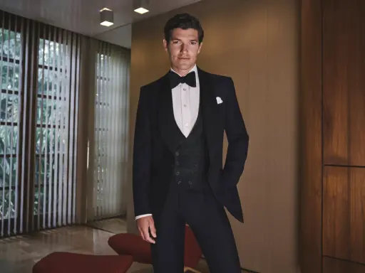 A man in a sharp black tie tuxedo, ready for a wedding. He stands confidently in a stylish room with large windows and wood paneling.