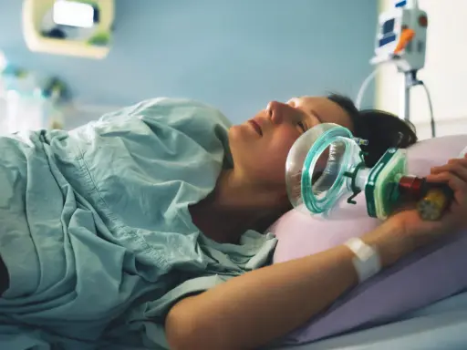 Kobieta w szpitalnym łóżku, z maską gazu rozweselającego przy twarzy, przygotowuje się do porodu.