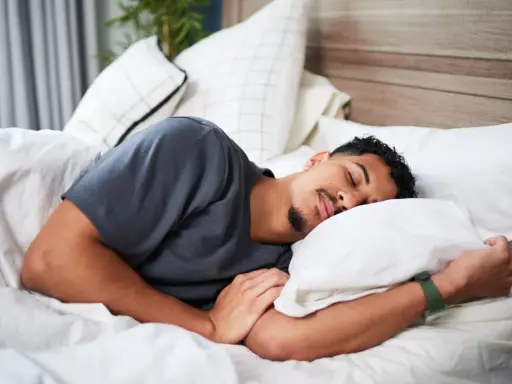 Ein Mann schläft tief und fest, um sein HRV zu verbessern. Er liegt auf einem weißen Bett mit vielen Kissen.