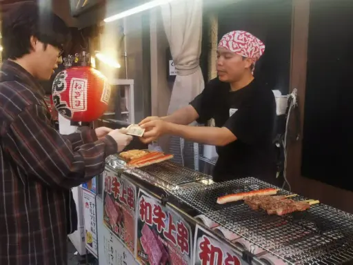 Un cliente paga por comida callejera japonesa: brochetas de carne y surimi a la parrilla en un puesto.