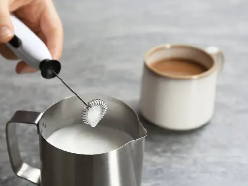 Hand hält Milchaufschäumer über Metallkännchen mit Milch. Perfekt für leckere Milchschaum Rezepte.