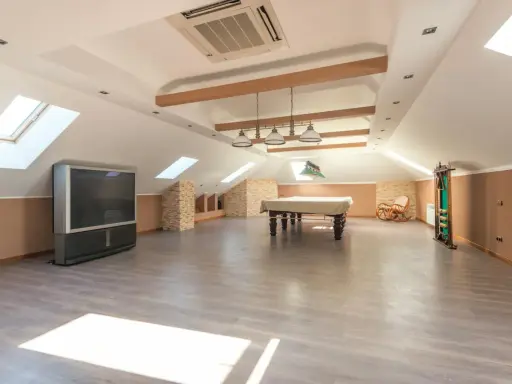 Vaste pièce sous les combles aménageables, avec table de billard, TV, fauteuil à bascule et étagères. Idéal pour un espace de loisirs.