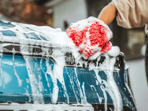 Mano con guante rojo y esponja enjabonada limpiando la carrocería del coche azul.
