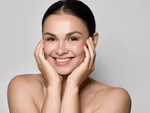 Uśmiechnięta kobieta z idealną, lśniącą cerą typu "glass skin", delikatnie dotykająca policzków dłońmi.